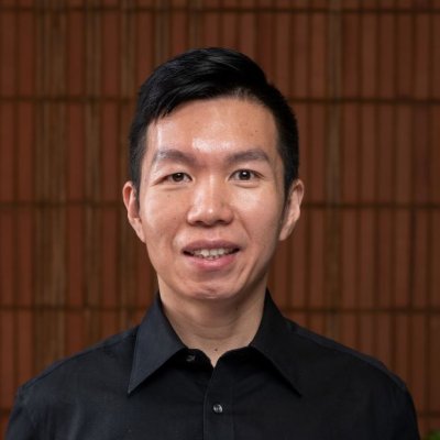 A man with black hair and wearing a black collared shirt stands in front of a brown timber wall. He is smiling at the camera.